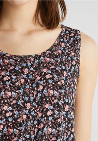 Gros plan sur un haut noir sans manches avec un petit motif floral rose et bleu porté par une personne ayant des cheveux bruns à longueur d'épaules.
