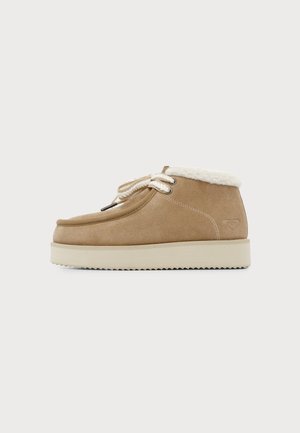 Chaussure beige en daim à la cheville avec doublure en polaire blanche, semelle épaisse blanche, bout rond et lacets blancs en corde sur un fond clair uni.