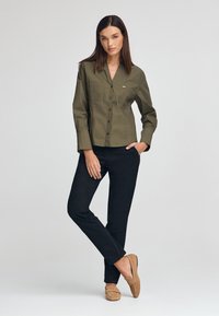 Olivgrön skjorta med knappar och bröstficka, kombinerad med svarta byxor och bruna loafers. Slät yta, strukturerad design, avslappnad passform.