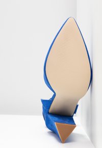 Chaussure à talon haut bleue vue de la semelle, bout pointu, embout de talon texturé, semelle beige, matériau en daim, positionnée contre une surface blanche.