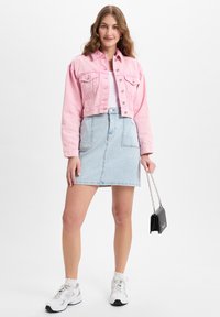 Rosa, kurzes Denim-Jackett mit zwei Brusttaschen, kombiniert mit einem hellblauen Denim-Rock und weißen Sneakers, hält eine schwarze Kettentasche.