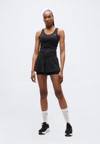 Γυναίκα που στέκεται με το σώμα στραμμένο προς τα εμπρός, φοράει μαύρο αθλητικό τοπ, μαύρα στρωμένα σορτς, λευκές κάλτσες μέχρι τον αστράγαλο και μαύρα αθλητικά παπούτσια σε απλό φόντο.