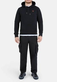 Zwarte pullover hoodie met een voorzak en trekkoorden. Gecombineerd met zwarte cargobroek met zijzakken. Draagt zwarte sneakers.