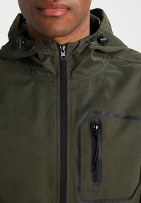 Detailní záběr osoby v olivově zelené kapuci s černým zipem a kapsou na hrudi se zipem a černým lemováním.