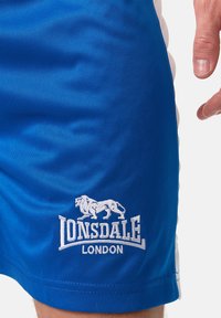 Mėlyni sportiniai šortai, pagaminti iš lygaus audinio, su balta siuvinėta liūto logotipu, po kuriuo užrašyta "LONSDALE" ir "LONDON".