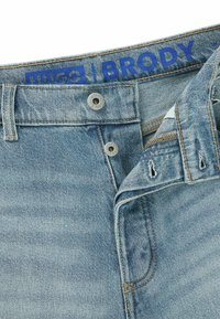 Jasnoniebieskie dżinsy z zagiętym pasem. Posiadają zapięcie na guzik, kontrastowe przeszycia oraz niebieską metkę marki na wewnętrznej stronie pasa.