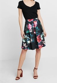 Černá květinová sukně s volánovým designem, zdobená barevnými růžemi a listy, spárovaná s černým žebrovaným topem a hnědými sandály na bloku.