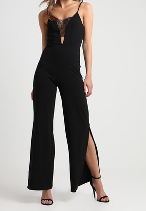 Mujer con mono negro sin mangas con escote en V de encaje y aberturas laterales, combinado con sandalias negras de tacón alto.