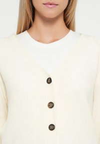 Cremefarbener Strickcardigan mit tiefem V-Ausschnitt, ausgestattet mit vier großen dunklen Knöpfen und einem gerippten Struktur an den Bündchen, über einem weißen Oberteil getragen.