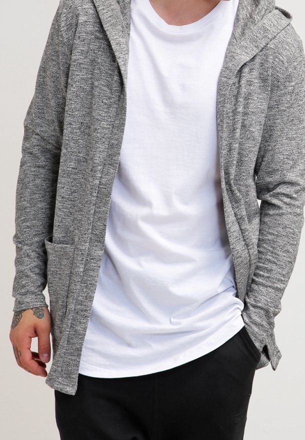 Persona che indossa un cardigan grigio con cappuccio sopra una maglietta bianca e pantaloni neri, con un tatuaggio visibile sulla mano sinistra.