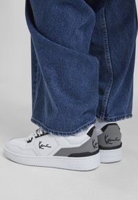 Baskets blanches en cuir avec des accents noirs et gris, design à bout rond et marque emblématique. Portées avec un pantalon en denim large et foncé.