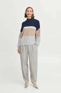 Pull en tricot à rayures bleu marine, beige et gris ; col montant ; coupe ample. Associé à un pantalon gris et des escarpins argentés à bout pointu.