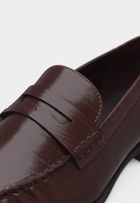 Καφέ δερμάτινη μοκασίνι με υφή αποτυπωμένη σαν κροκόδειλος και κλασική λεπτομέρεια με λουράκι μπροστά, που δείχνει λεπτομερή ραφή και λείο φινίρισμα.