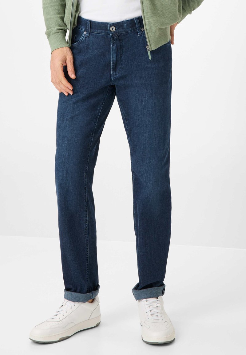 Dark blue denim jeans with straight cut, featuring a button and zipper closure, front pockets, and rolled cuffs. Worn with white sneakers.