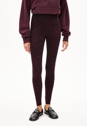 Burgunderfarbene Leggings mit einem schwarzen Zebramuster, aus dehnbarem Stoff, kombiniert mit schwarzen Slippern. Hohe Taille.