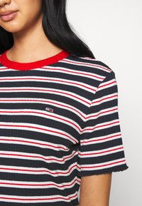 Czarny i biały pasiaste, ribbowe t-shirt z czerwonymi akcentami i falbaniastym dołem. Małe logo na piersi. Krótkie rękawy i okrągły dekolt.