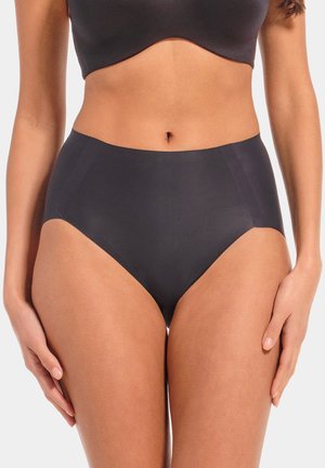 Femme portant des sous-vêtements noirs sans coutures taille haute et un crop top noir, debout les mains détendues le long du corps sur un fond neutre.