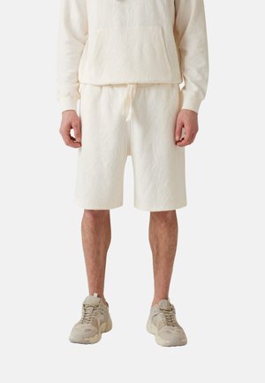 Sudadera beige claro y pantalones cortos a juego, hechos de una tela texturizada con patrones de hojas. Combinados con zapatillas de tonalidad neutra.
