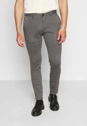 Mężczyzna ubrany w szare spodnie typu slim fit, czarne buty sznurowane i białą koszulę włożoną do spodni, stojący na jednolitym jasnym tle.
