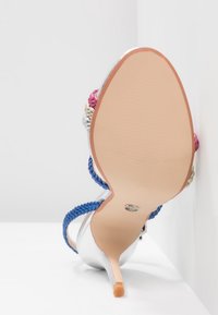 Chaussure à talon haut argentée avec des brides bleues tressées, des accents floraux en rose et crème, et une semelle lisse couleur nude.