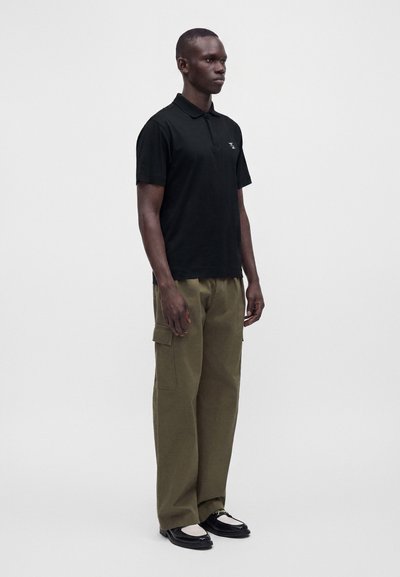 Homme debout face à nous, portant un polo noir, un pantalon cargo olive et des chaussures slip-on noires avec un détail blanc.
