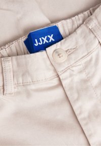 Krémové bavlněné kalhoty s modrou nášivkou "JJXX", zapínání na knoflík a hladkou texturu. Obsahují detaily šití poblíž pasu.