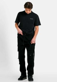 Zwarte t-shirt met korte mouwen en een klein logo, gecombineerd met zwarte cargobroeken met meerdere zakken en ritsen. Sneakers maken de look compleet.