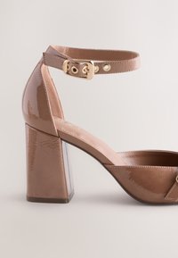 Sandalo con tacco alto in pelle verniciata beige, tacco largo, cinturino alla caviglia, fibbia dorata e punta arrotondata. Texture liscia con dettagli di design minimalisti.