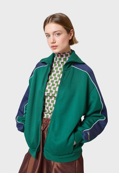 Zelena i tamnoplava jakna na zip s bijelim obrubom, koja ima visoki ovratnik i široku kroj, upotpunjena uzorkovanom, prilagodenom majicom ispod.