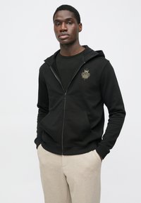Čierna mikina na zips z mäkkej látky s predným vreckom a zlatým logom na ľavej strane hrudníka. Nosená cez čierne tričko.