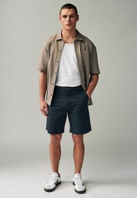 Camisa corta de manga beis con botones, camiseta blanca, pantalones cortos azul marino y zapatillas blancas con detalles en negro. Diseño sencillo y ajuste casual.