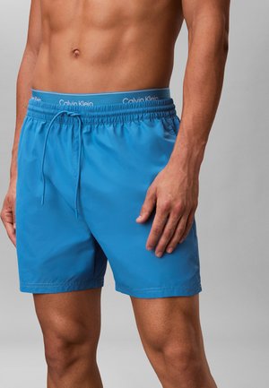 Männlicher Torso trägt blaue Badeshorts mit elastischem Bund und Kordelzug, sichtbarer Bund der Calvin Klein Unterwäsche oberhalb der Shorts.
