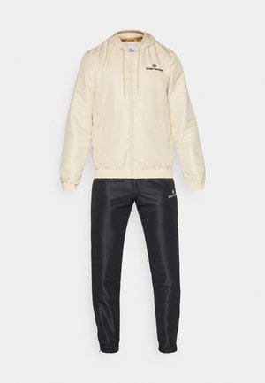 Chaqueta de satén crema con capucha, con puños elásticos y un logo; combinada con pantalones jogger de satén negros con puños elásticos en el tobillo y detalles del logo.