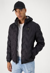 Giacca a vento nera con cappuccio, caratterizzata da un motivo a losanga trapuntato e chiusura con zip. Indossata sopra una maglietta bianca e abbinata a jeans chiari.