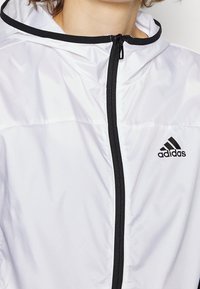 Bílá lehká bunda s černým zipem a kapucí, s logem Adidas. Hladká textura s čistým, minimalistickým designem.