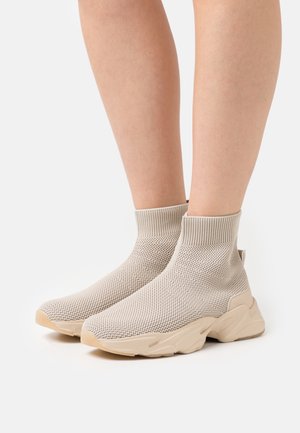 Piernas con zapatillas deportivas de calcetín tejidas en beige y suelas gruesas, de pie sobre una superficie blanca.