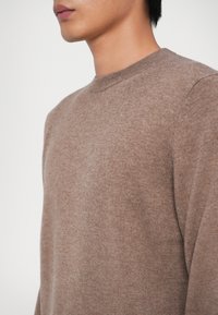 Brun strikket sweater med rund hals, glat overflade og forlængerede ærmer. Stoffet ser blødt og let tekstureret ud.