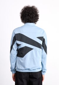 Light blue zip-up jacket with black geometric design elements, ribbed cuffs and waistband, and smooth texture. Suitable for casual wear.
