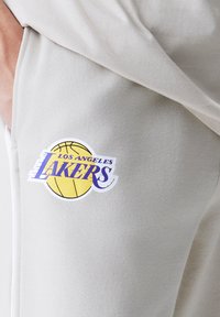 Calças de moletom cinzas com o logótipo dos Los Angeles Lakers, apresentando um design de basquetebol e acabamentos em roxo, amarelo e branco num tecido suave.