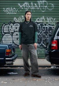 Zelené tričko s potiskem vrstvené přes tričko s dlouhým rukávem, doplněné volnými šedými džíny a hnědými dřeváky, na pozadí stěny pokryté graffiti.