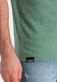 Camiseta de algodón verde con acentos amarillos moteados, con una etiqueta negra en el dobladillo. Combinada con pantalones vaqueros desgastados de un azul claro.