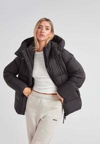 Czarna kurtka puchowa z kapturem, pikowane wzornictwo, zatrzaski i luźny krój, noszona na białej bluzce i beżowych dresach.