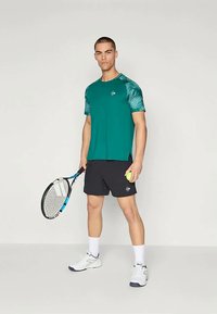 Žalias trumpomis rankovėmis marškinėliai su raštuotomis rankovėmis, derinami su juodomis šortais. Modelis laiko teniso raketę ir kamuoliuką, vilkėdamas baltus sportbačius.