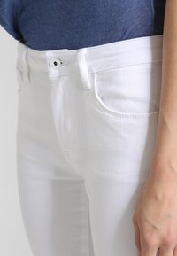 Jeans di denim bianco con una texture liscia, design a vita alta e stile classico a cinque tasche. Presentano una chiusura a bottone anteriore.