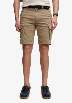 Mand iført beige cargoshorts, sort flettet bælte, sort ur og sorte bådsko, der står med armene ned langs siderne.