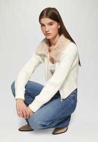 Weißer gerippter Cardigan mit einem weichen beigen Pelzkragen, kombiniert mit blauen Jeans und beigen Ballerinas, in einer hockenden Pose vor einem hellen Hintergrund dargestellt.