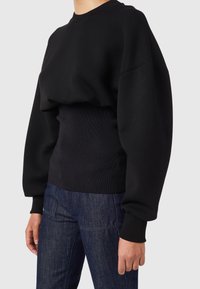Černá mikina s volným střihem, žebrovaným pasem a volnými rukávy. Vyrobena z měkkého materiálu s minimem švů a hladkou texturou.