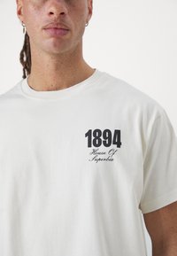 White cotton t-shirt with round neckline. Front features black printed text: "1894" and "House Of Superbia." Smooth texture, casual fit.
