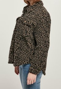 Kobieta w brązowo-czarną kurtkę w leopardzi wzór z teksturą, z przednimi kieszeniami i niebieskimi dżinsami, pokazana z boku.