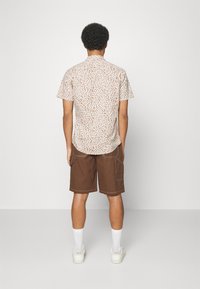 Camisa de manga corta con un estampado floral en tonos marrón claro y blanco, combinada con pantalones cortos marrones que cuentan con bolsillos y costuras contrastantes.